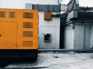 Outdoor view of an industrial generator with visible piping and wiring against a concrete wall.